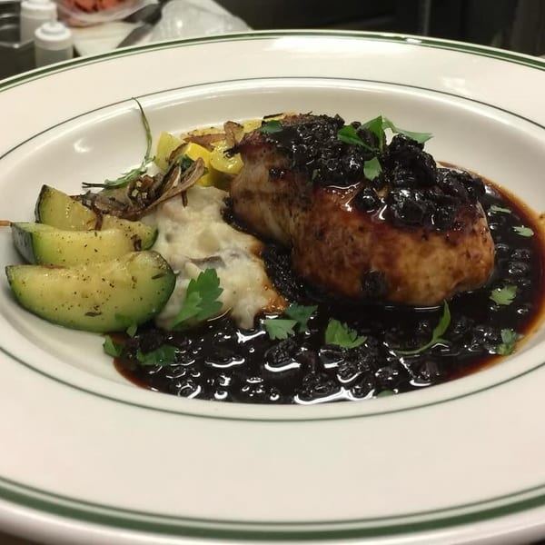Pork chop with dark berry sauce, mashed potatoes, and zucchini on a white plate.