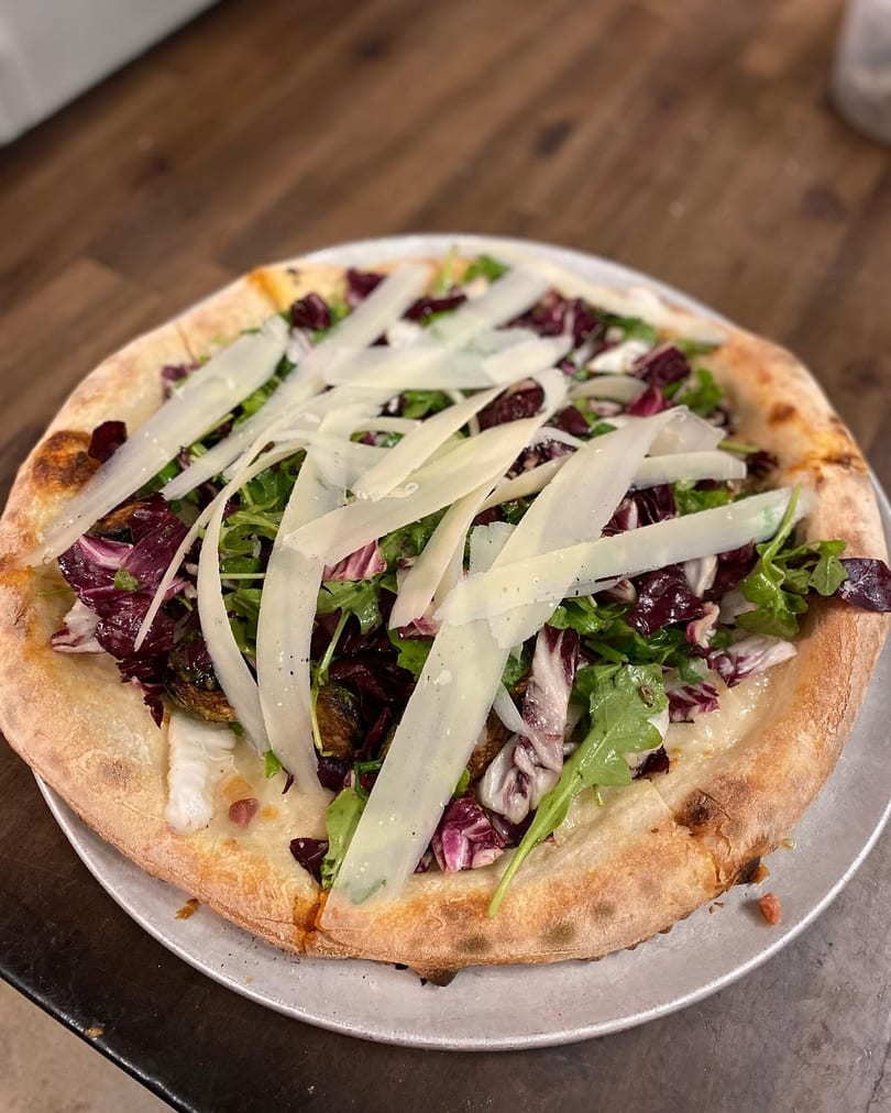 A pizza topped with fresh arugula, radicchio, roasted Brussels sprouts, and shaved Parmesan cheese.