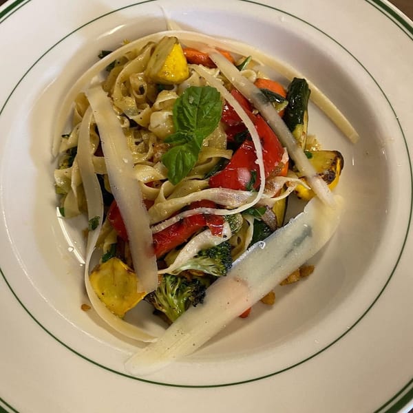 Fettuccine pasta with grilled vegetables, parmesan shavings, and a fresh basil leaf in a white bowl.