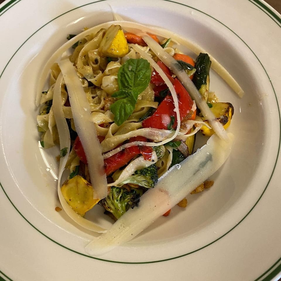 Fettuccine pasta with grilled vegetables, parmesan shavings, and a fresh basil leaf in a white bowl.