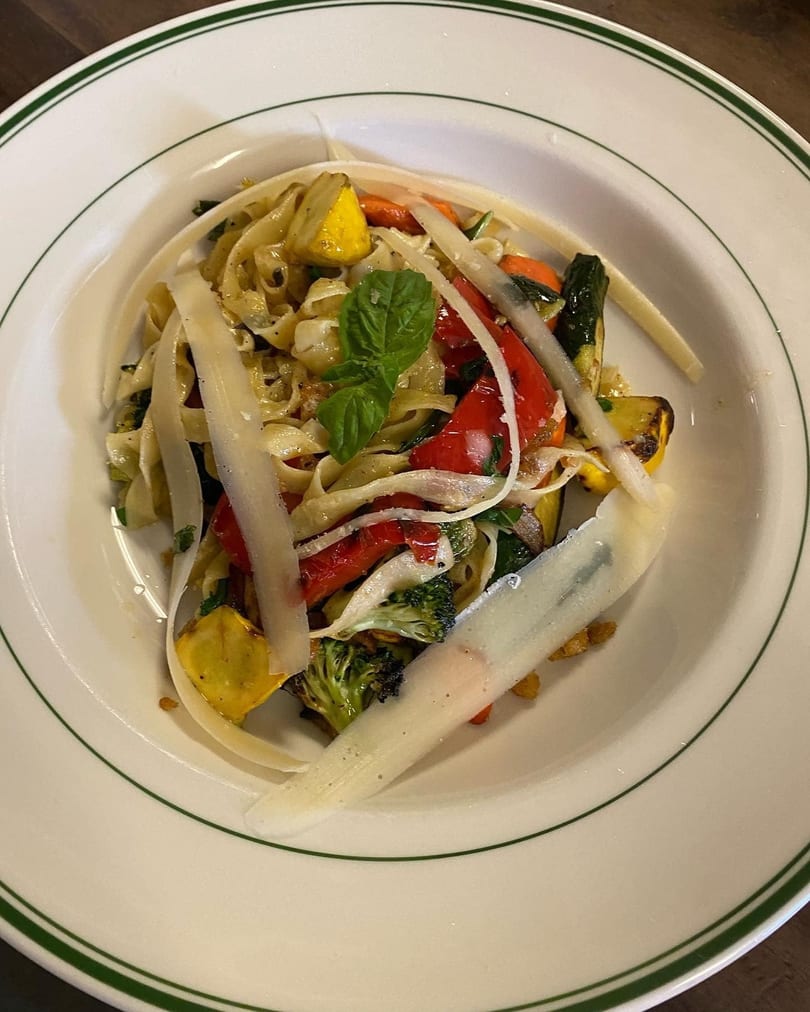 Fettuccine pasta with grilled vegetables, parmesan shavings, and a fresh basil leaf in a white bowl.