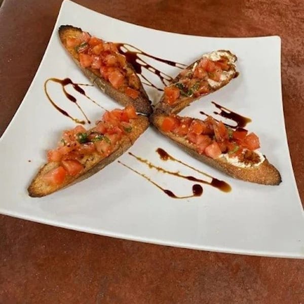 Four bruschetta pieces with diced tomatoes and balsamic glaze on a white square plate.