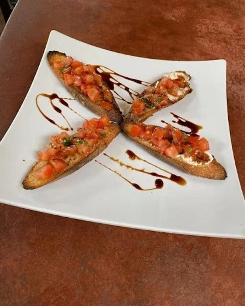 Four bruschetta pieces with diced tomatoes and balsamic glaze on a white square plate.